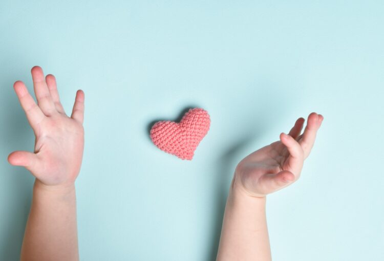 Children's hands hold a small pink knitted heart on a blue background.